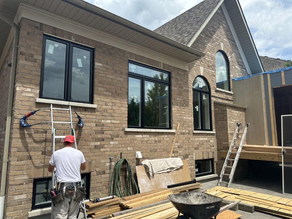 Installer on a ladder beside new black-frame windows on a brick house