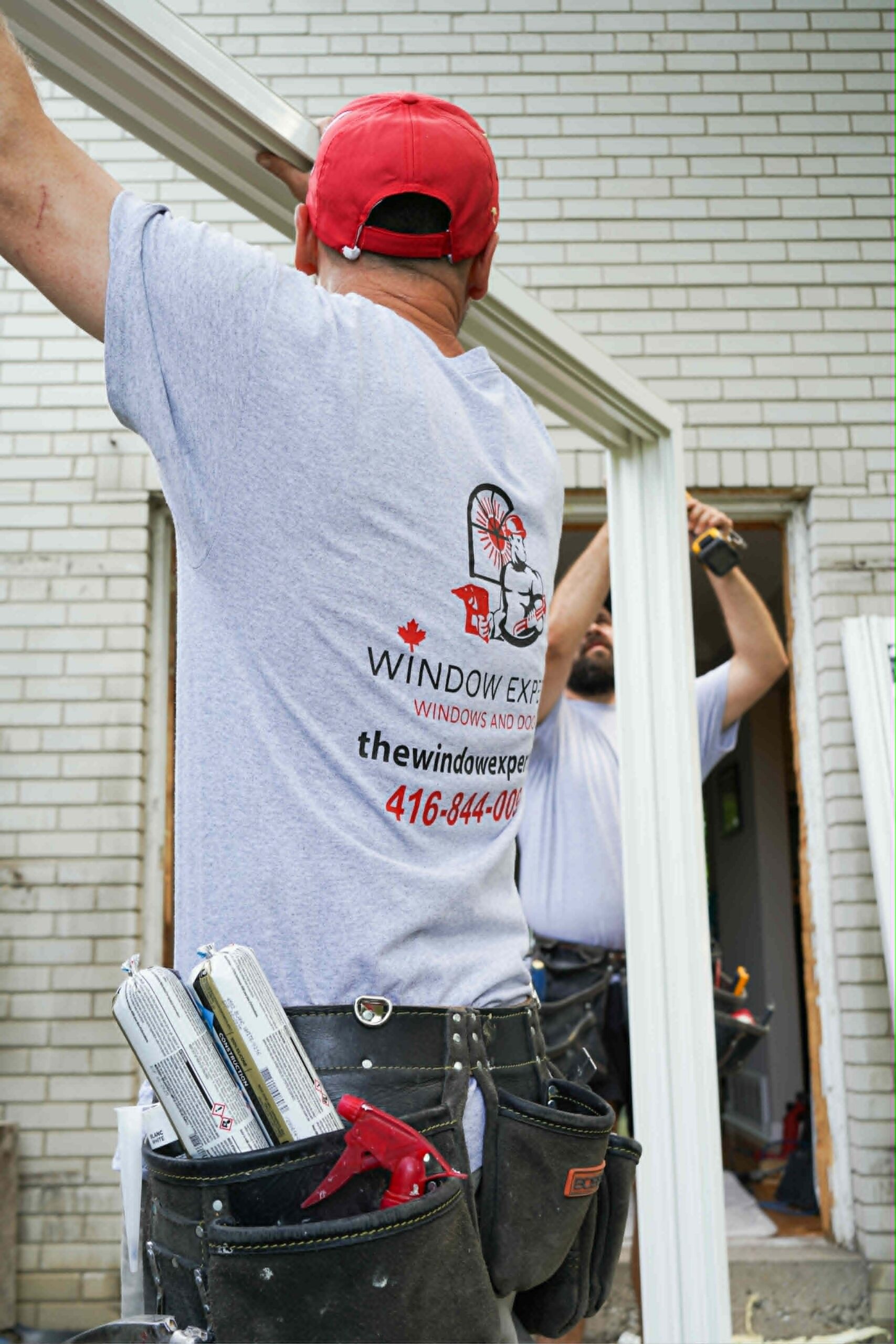 Two Window Experts installers in branded shirts and tool belts fitting a white window frame into a brick home