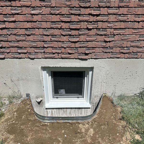 Newly installed basement egress window in concrete foundation below brick wall with metal window well and dirt
