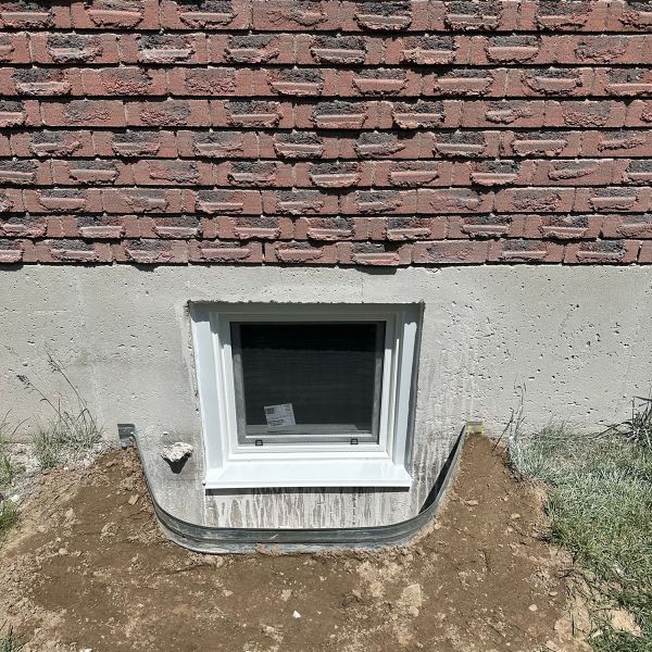 Newly installed basement egress window in concrete foundation below brick wall with metal window well and dirt
